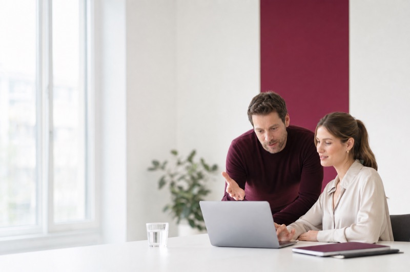 Zwei Kollegen besprechen sich an einem Laptop in einem hellen Büro